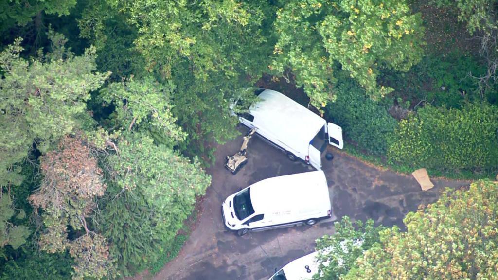 Aerial still of three white vans on a tree-lined lane in Whelford, two with rear doors open, and an Army bomb-disposal robot between them